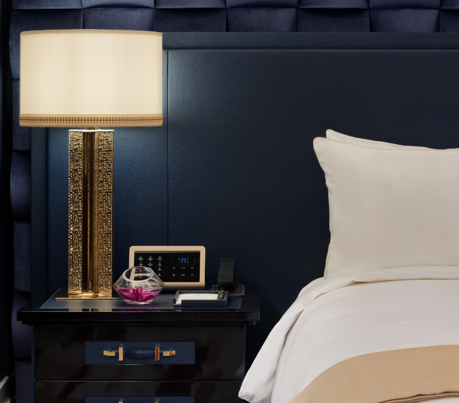 Corner of a bed with an end table with a lamp, phone and alarm clock