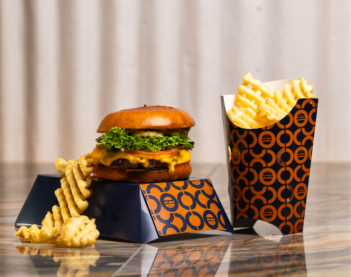 Hamburger with lettuce, tomato, cheese and buns, cup of waffle cut french fries with a few fries next to the burger