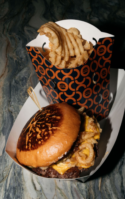 Hamburger with lettuce, tomato, cheese and buns, cup of waffle cut french fries with a few fries next to the burger