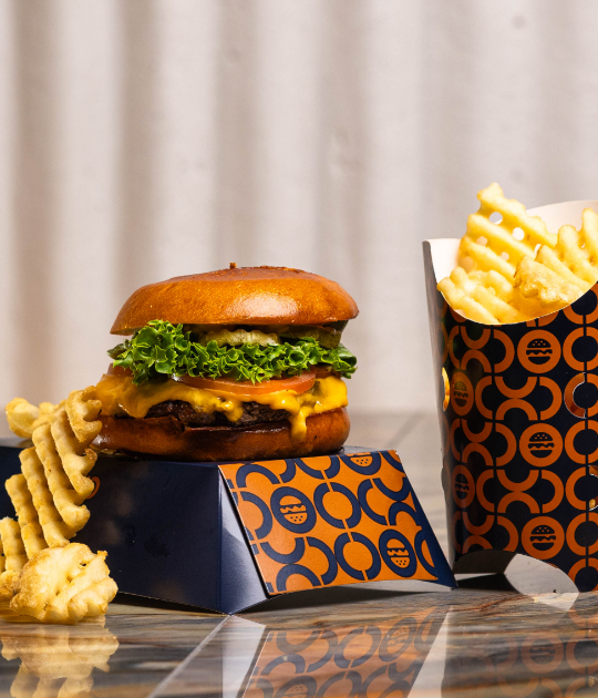 Hamburger with lettuce, tomato, cheese and buns, cup of waffle cut french fries with a few fries next to the burger