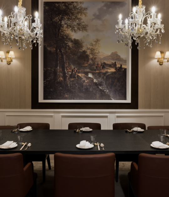 6-person black dining table with brown chairs and place settings underneath 2 chandeliers with a painting on the back wall surrounded by 2 sconces