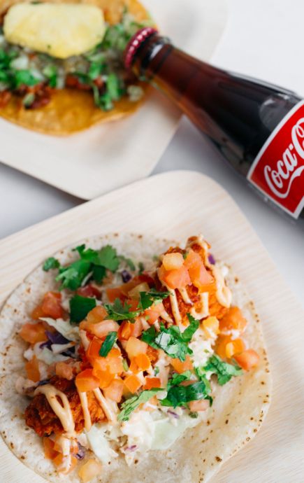 Filled taco laying open on a surface with a bottle of Coca-Cola laying on its side with a second taco in the background