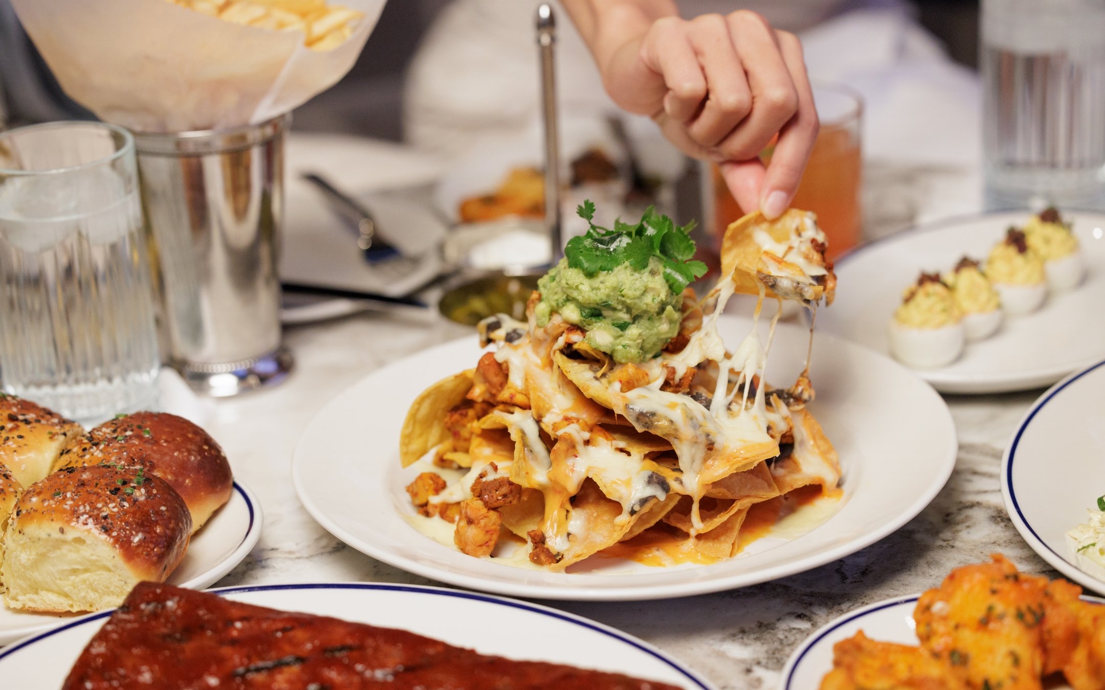 Hand pulling a nacho away from a pile of nachos with cheese stretching between them, and surrounded by deviled eggs, buns and other food items