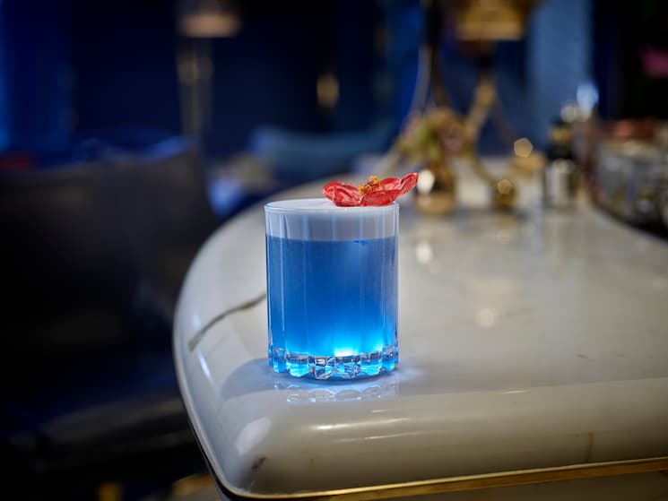 Blue cocktail with red flower garnish on white granite countertop