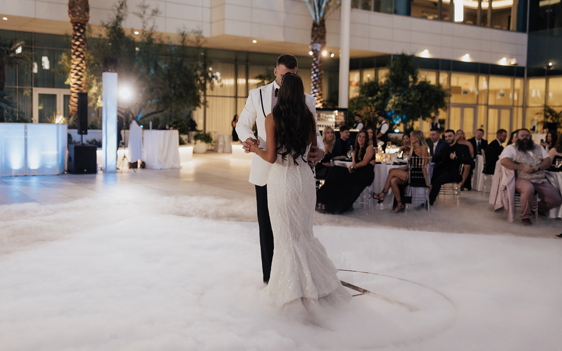 Couple dancing on a dance floor in wedding outfits with a crowd sitting around