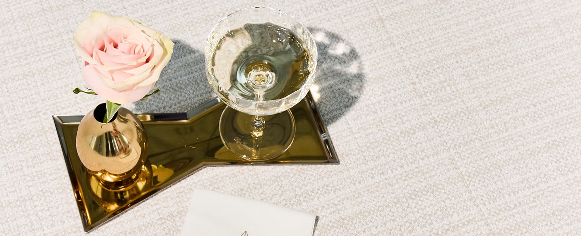 Gold bowtie tray with a pink rose in a gold vase next to a champagne glass on a beige tweed background