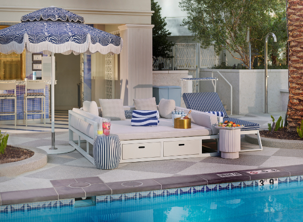 Daybed with lounge chair, umbrella, towels, bottles, champagne, fruit tray, next to a pool