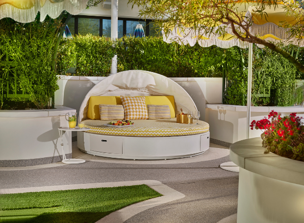 Daybed with lounge chair, umbrella, towels, bottles, champagne, fruit tray with yellow accents near foliage