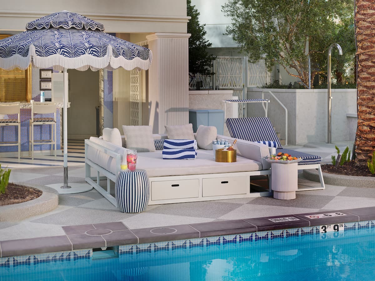 Daybed with lounge chair, umbrella, towels, bottles, champagne, fruit tray, next to a pool