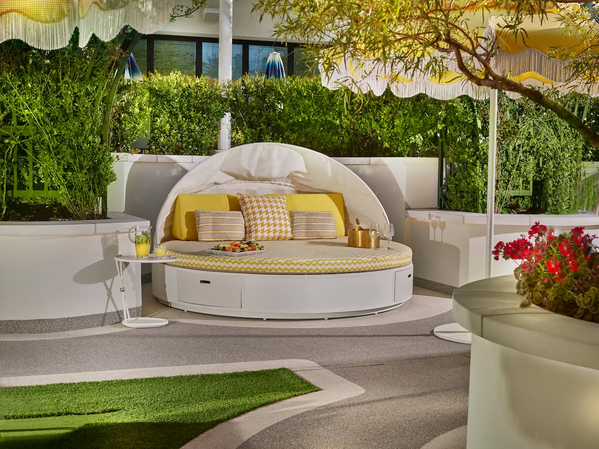 Daybed with lounge chair, umbrella, towels, bottles, champagne, fruit tray with yellow accents near foliage