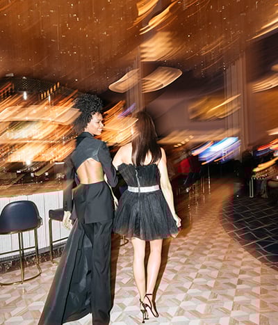 Pair of women in evening dresses walking through a deliberately blurred background in Bleau Bar