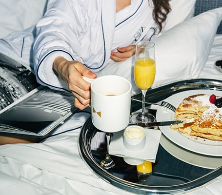 Person in a robe with a cup of coffee reaching towards a tray of pancakes and mimosa for room service at Fontainebleau Las Vegas