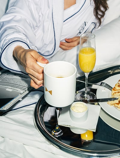 Person in a robe with a cup of coffee reaching towards a tray of pancakes and mimosa for room service at Fontainebleau Las Vegas
