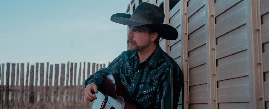 Mustached man with cowboy hat and guitar against a wall