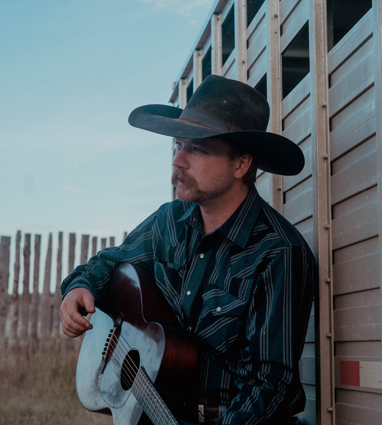 Mustached man with cowboy hat and guitar against a wall