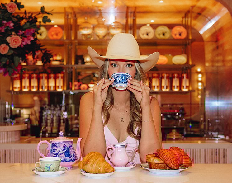 Woman in cowboy hat sipping out of a tea cup with pinkies raised over pastries and tea settings