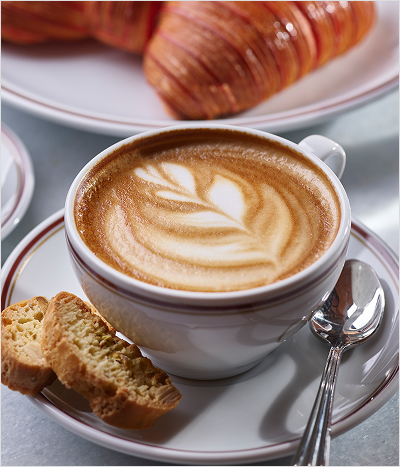 Latte with flowered design sitting on plate with bread and spoon