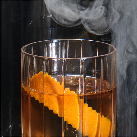 Glass of amber liquid with orange garnish with smoke