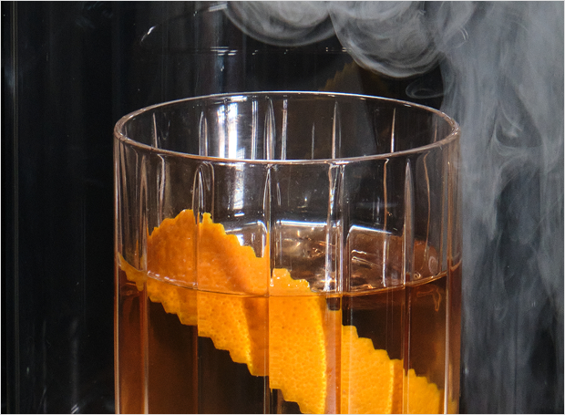 Glass of amber liquid with orange garnish with smoke