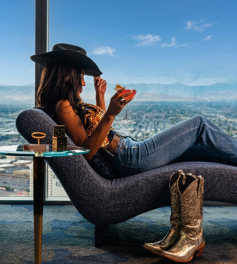 Cowgirl with designer black country western cowboy hat gold boots and jeans relaxing during NFR holding a pink cocktail sitting in purple lounge chair overlooking the Las Vegas Strip with view of the mountains and city with gold casino dice on glass display table next to her
